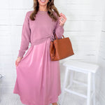 Woman wearing a pink dress with sunglasses and holding a brown bag against a white background