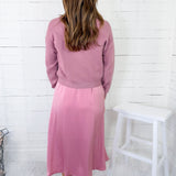 Woman wearing a pink outfit standing against a white wooden paneled wall.