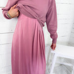 Person wearing a pink dress with long sleeves against a white wooden panel background