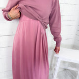 Person wearing a pink dress with long sleeves against a white wooden panel background