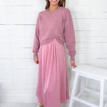 Woman wearing a pink sweater and pink skirt standing against a white wooden paneled wall.