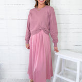 Woman wearing a pink sweater and pink skirt standing against a white wooden paneled wall.