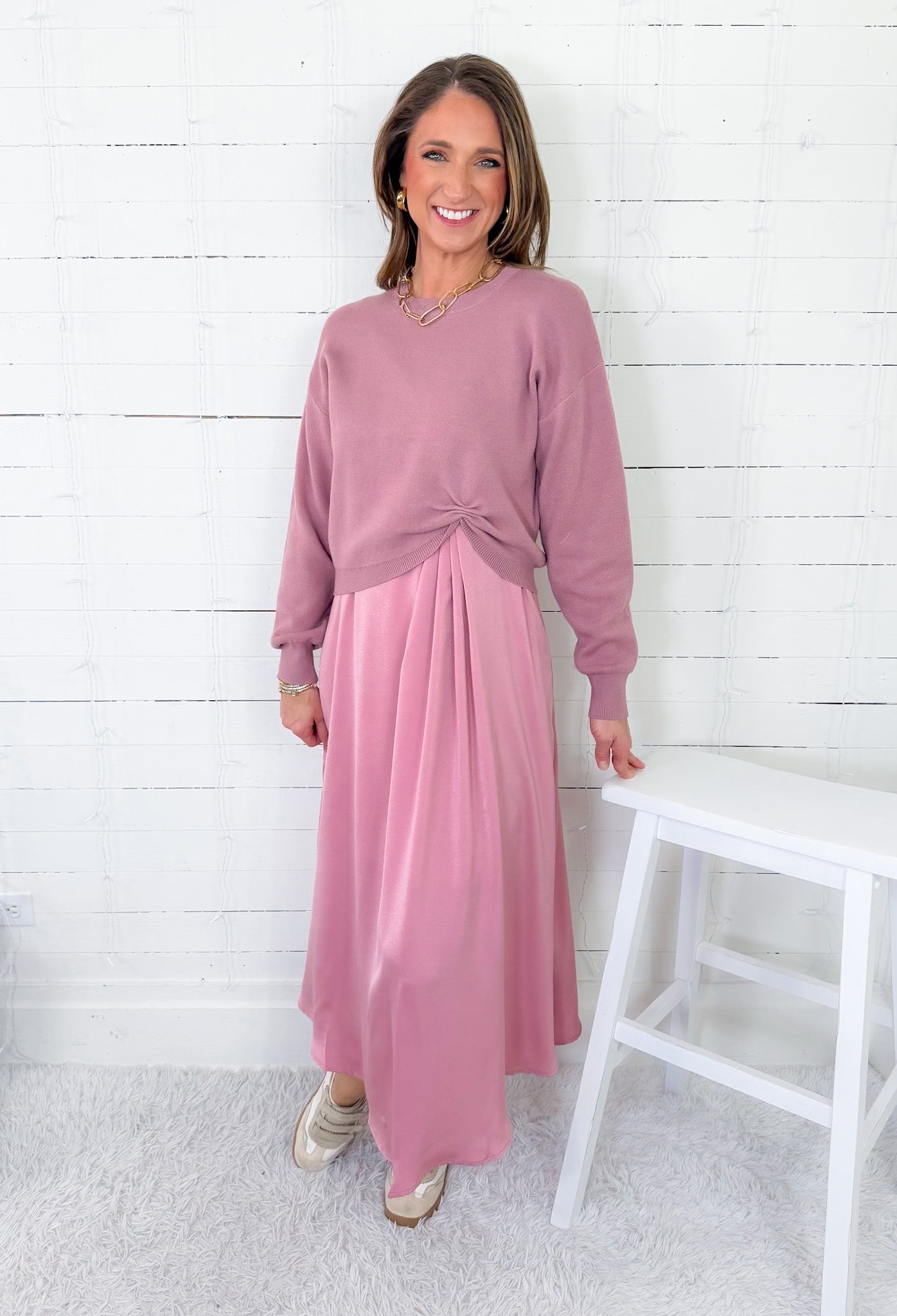 Woman wearing a pink sweater and pink skirt standing against a white wooden paneled wall.