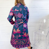 Woman wearing a long, floral-patterned dress with a blue base color against a white wooden panel background.