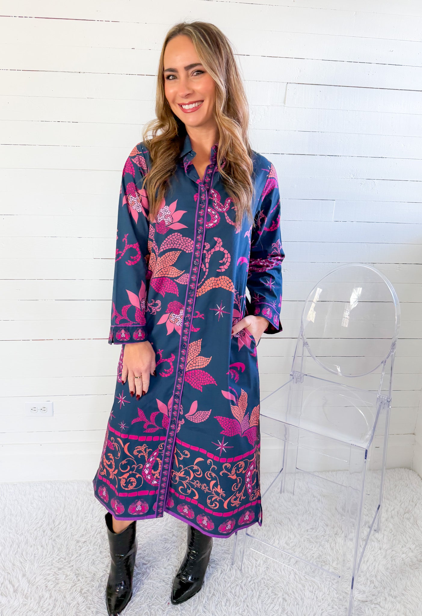 Woman wearing a blue and pink floral dress with black boots in front of a white wooden panel wall.