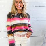 Woman wearing a colorful striped sweater and white pants against a white wooden panel background