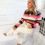 Woman sitting on a clear chair wearing a colorful striped sweater and white pants in a room with white walls.
