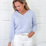Woman wearing a light blue sweater and white pants against a white wooden panel background