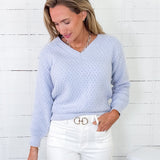 Woman wearing a light blue sweater and white pants against a white wooden panel background