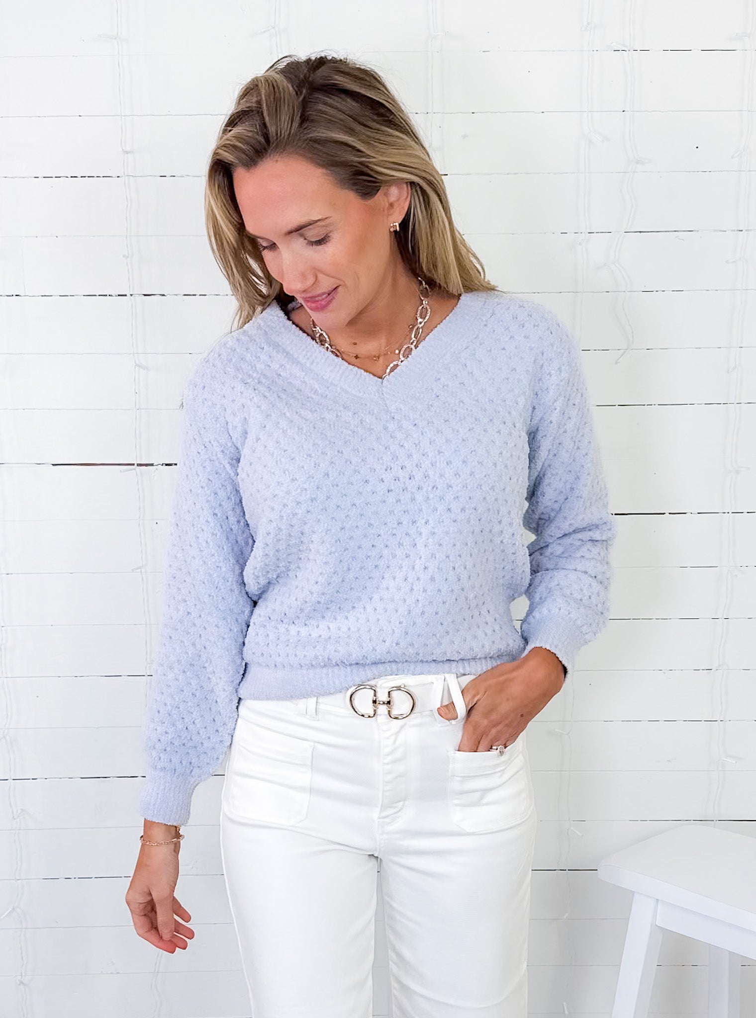 Woman wearing a light blue sweater and white pants against a white wooden panel background