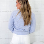 Woman wearing a light blue textured sweater and white pants against a white wooden panel background