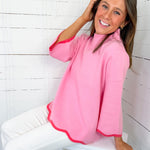Woman wearing a pink top with white pants sitting on a white stool against a white wooden paneled wall.