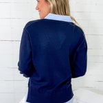 Woman wearing a navy sweater over a light blue shirt with white pants against a white wooden panel background