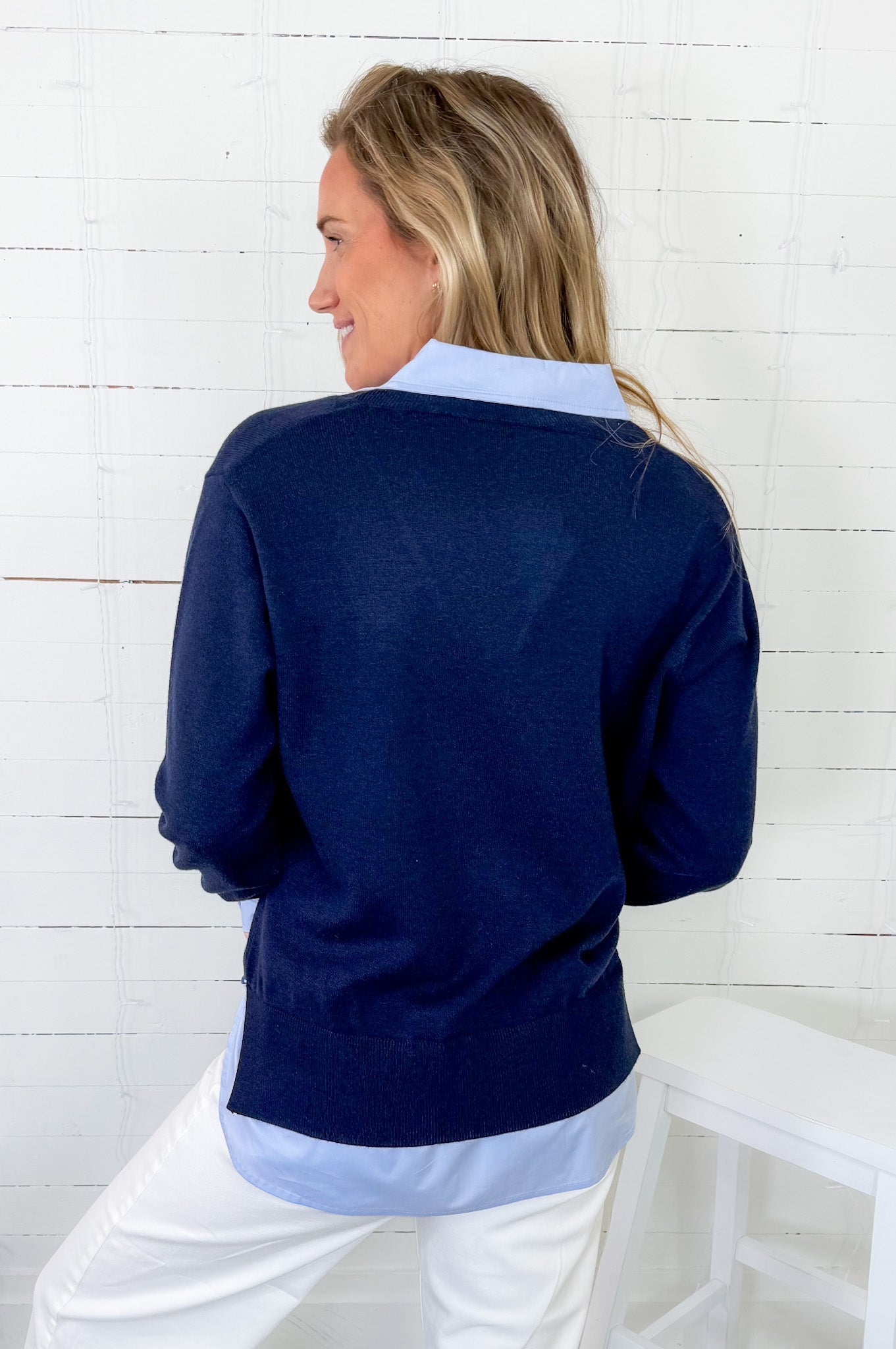 Woman wearing a navy sweater over a light blue shirt with white pants against a white wooden panel background