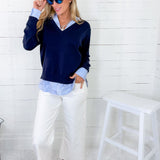 Woman wearing a navy sweater over a light blue shirt with white pants, standing against a white brick wall.