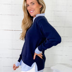 Woman wearing a navy sweater over a light blue shirt with white pants against a white wooden paneled wall.