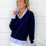 Woman wearing a navy sweater over a light blue shirt with white pants against a white wooden wall.