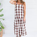 A woman modeling a brown and cream buffalo plaid midi dress with a tiered skirt, standing in front of a white wood-paneled wall next to a potted plant.