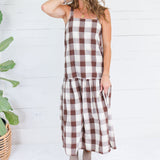 A woman modeling a brown and cream buffalo plaid midi dress with a tiered skirt, standing in front of a white wood-paneled wall next to a potted plant.