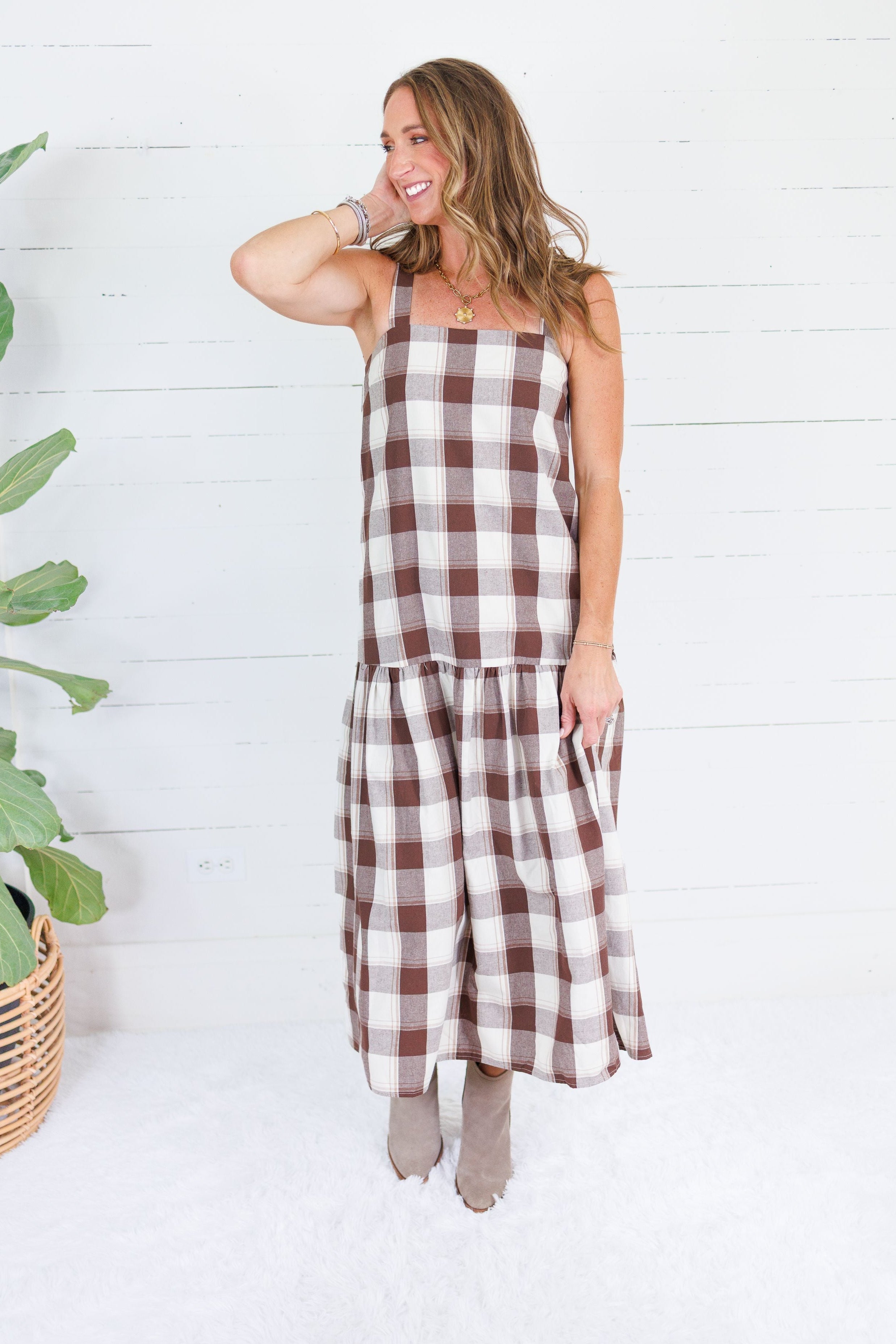 A woman modeling a brown and cream buffalo plaid midi dress with a tiered skirt, standing in front of a white wood-paneled wall next to a potted plant.
