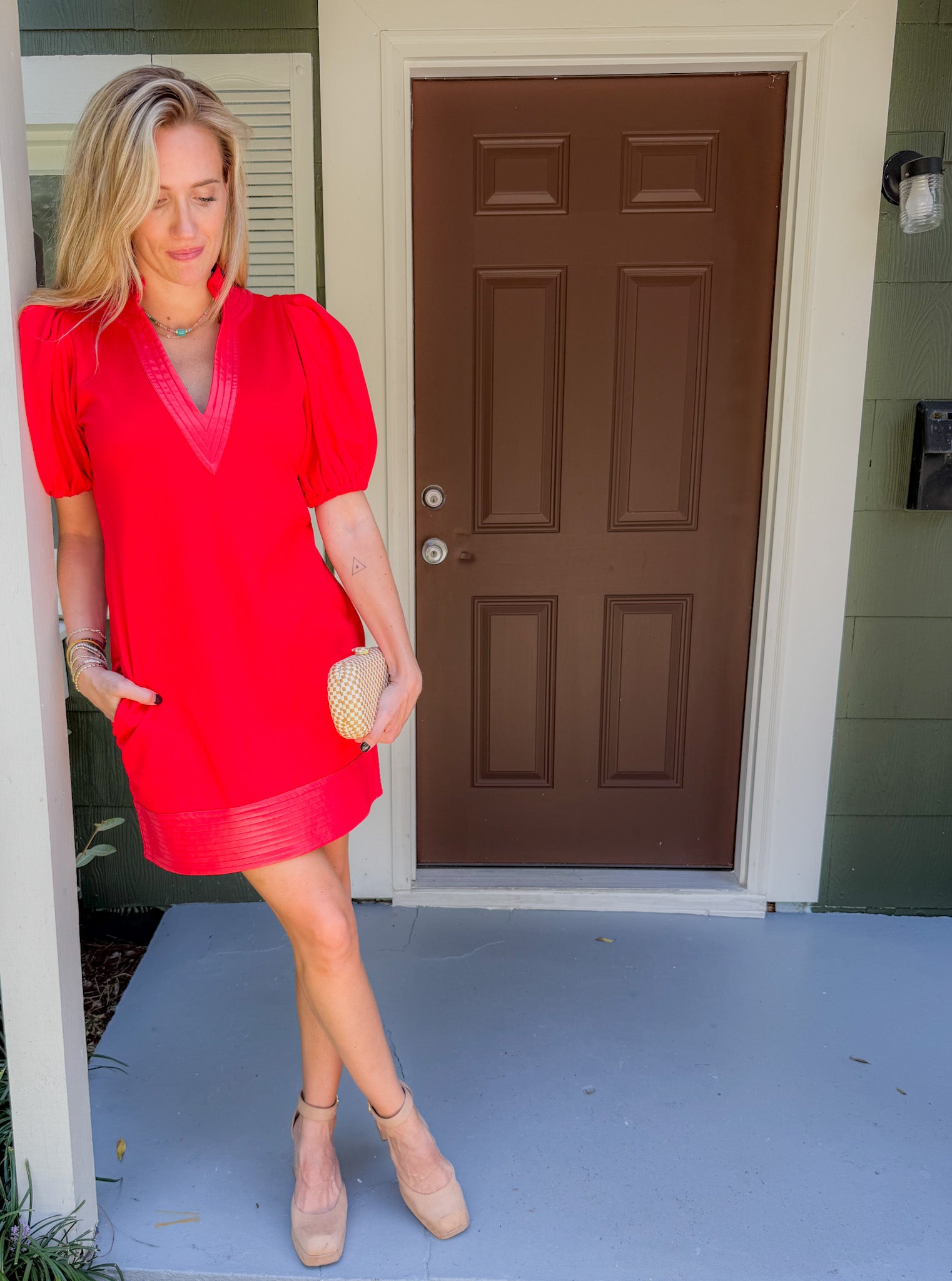 Woman in a red dress standing on a porch with a brown door.