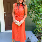 Woman in an orange dress standing on a porch with a brown door and greenery.