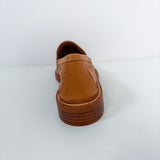 A single tan leather slip-on shoe shown from the back, featuring a thick brown platform sole with visible stitching, set against a plain white background.