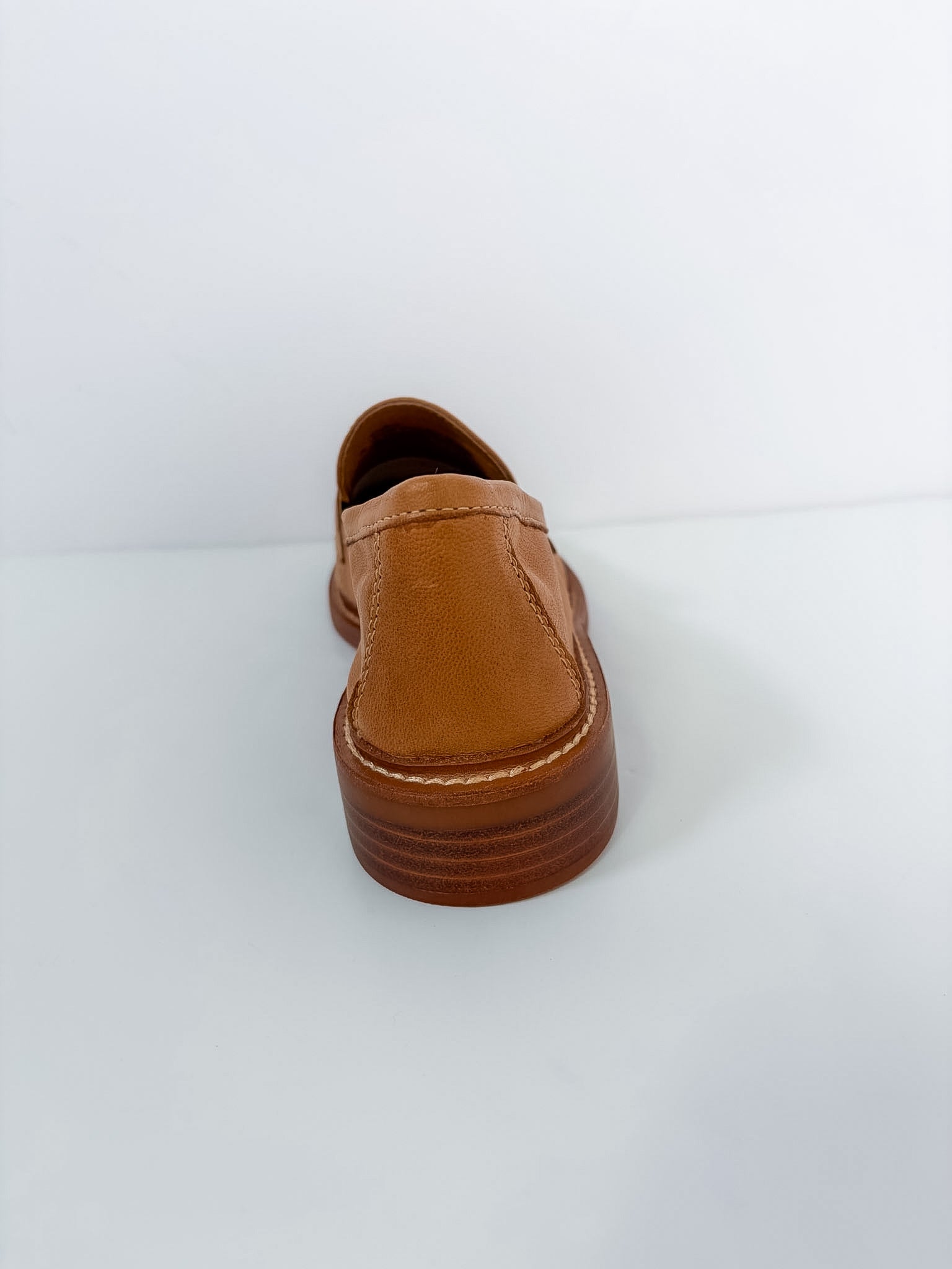 A single tan leather slip-on shoe shown from the back, featuring a thick brown platform sole with visible stitching, set against a plain white background.