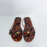 A pair of Versace flat sandals with a glossy tortoiseshell pattern in brown and amber, featuring thin brown straps and a small gold-tone circular medallion on the toe strap. The brand name 'VERSACE' is visible on the insole.