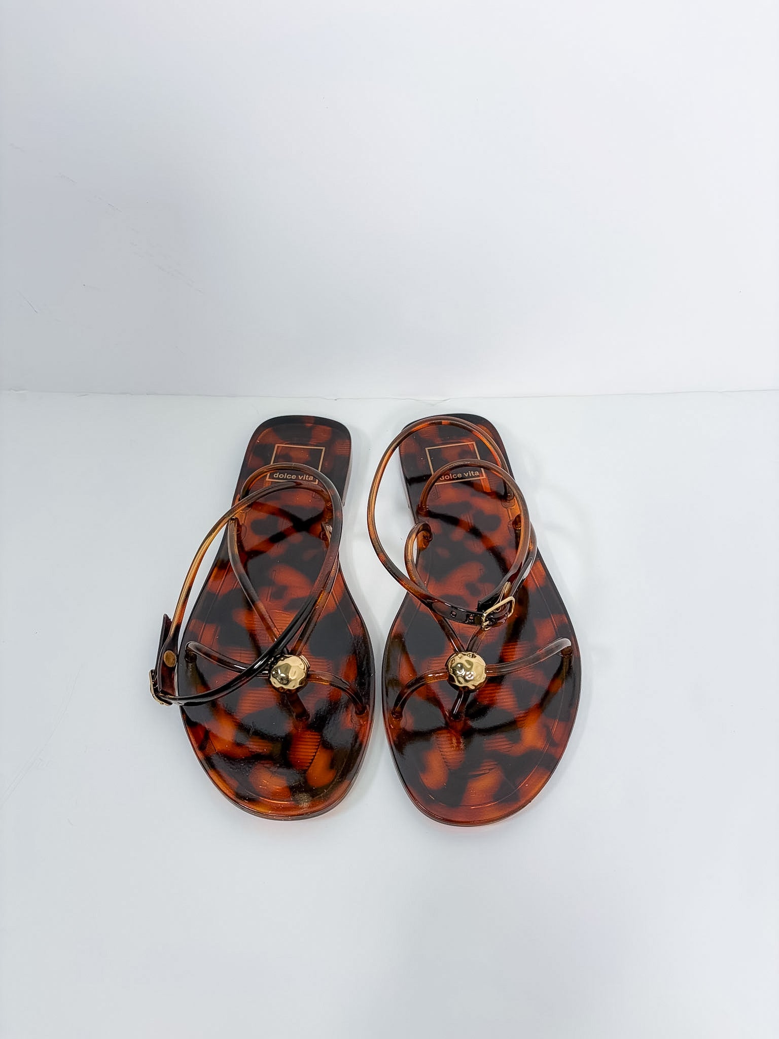 A pair of Versace flat sandals with a glossy tortoiseshell pattern in brown and amber, featuring thin brown straps and a small gold-tone circular medallion on the toe strap. The brand name 'VERSACE' is visible on the insole.