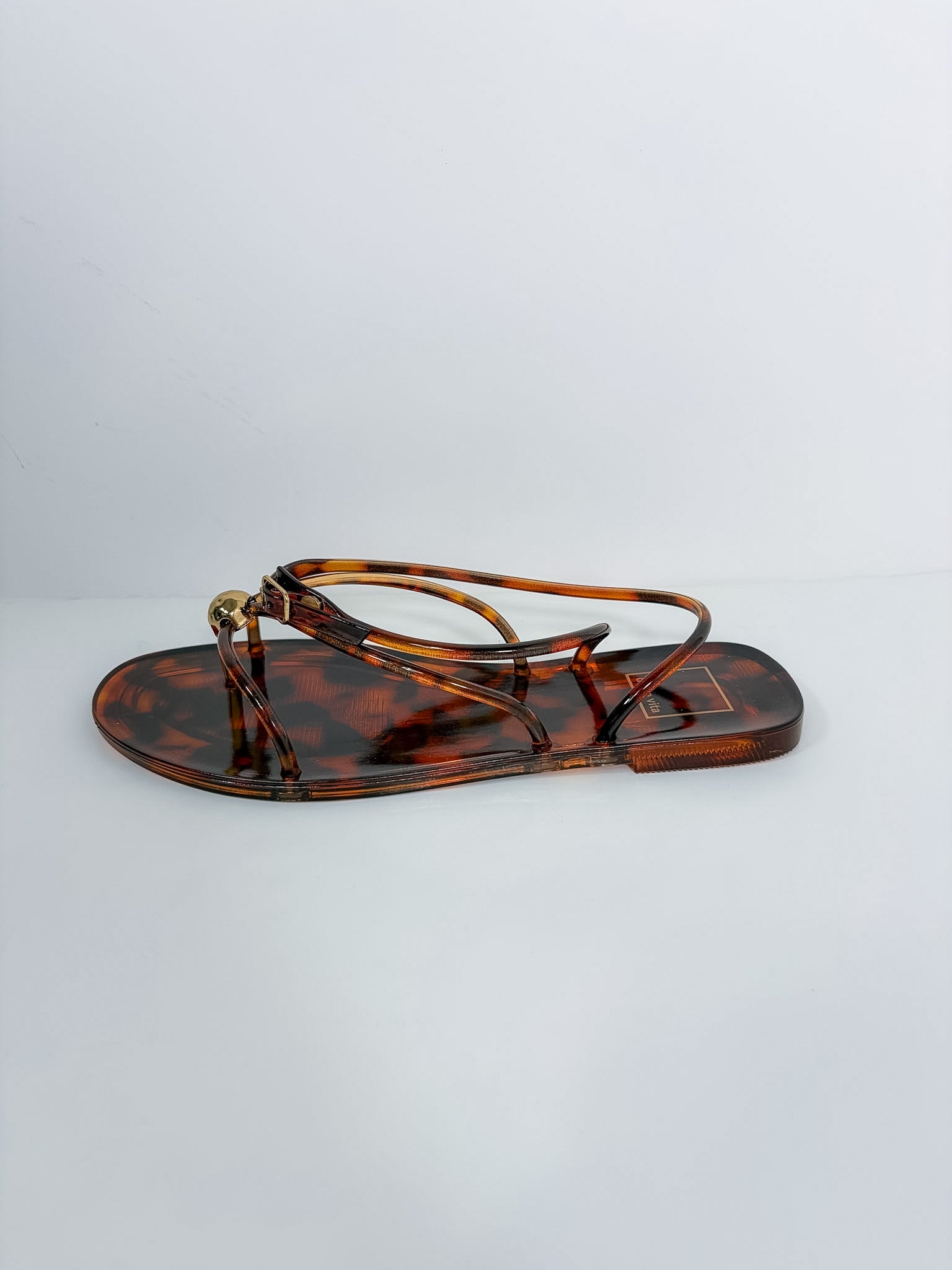 A single flat thong sandal with a glossy tortoiseshell pattern in brown and amber, featuring thin wrap-around straps and a small gold-tone metal ring detail at the front, shown against a plain light gray background.