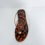 A single flat thong sandal with a glossy tortoiseshell pattern in brown and amber, featuring thin brown straps and a small gold-tone circular charm, photographed from a top-down angle on a white background.