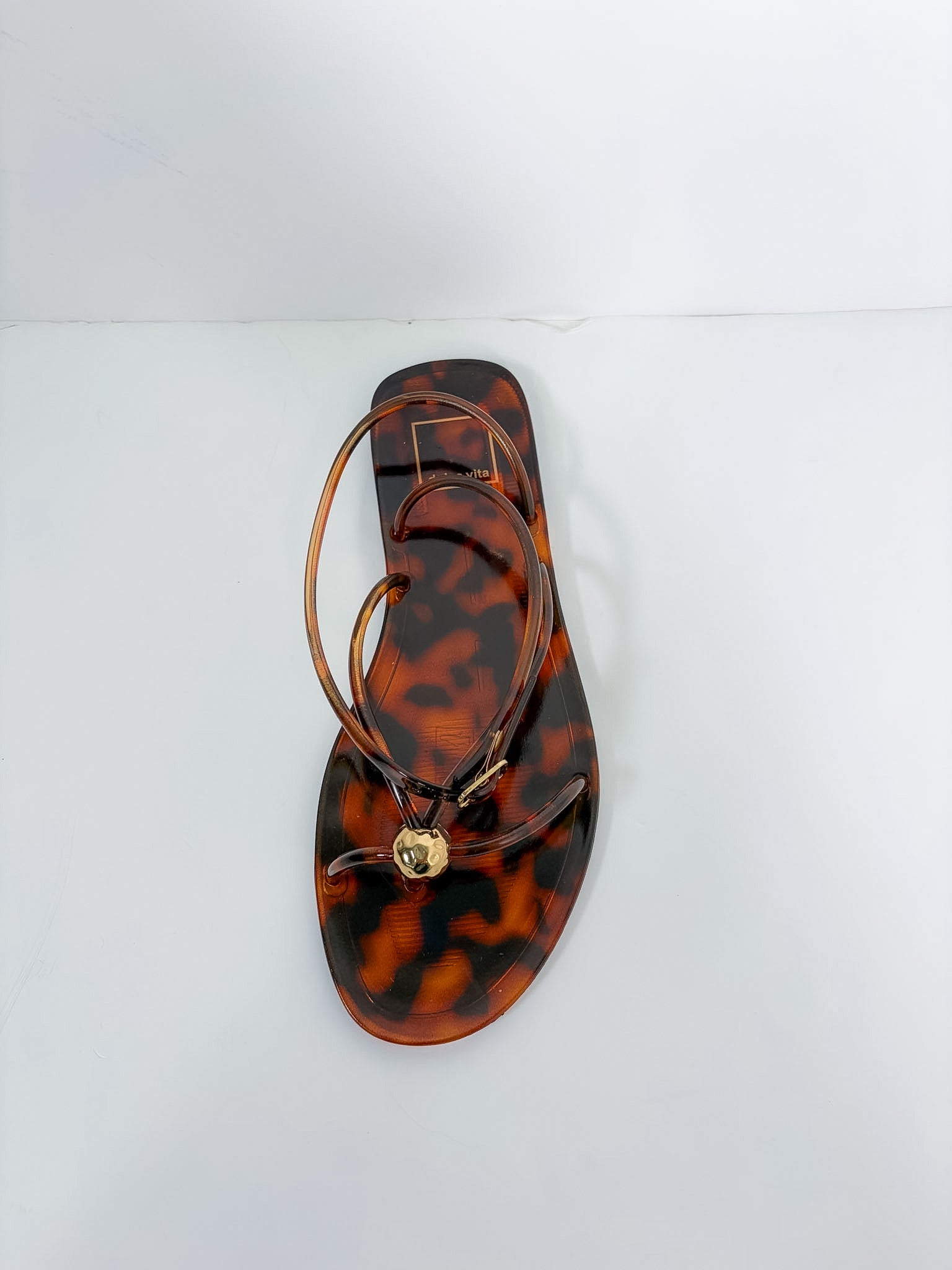 A single flat thong sandal with a glossy tortoiseshell pattern in brown and amber, featuring thin brown straps and a small gold-tone circular charm, photographed from a top-down angle on a white background.