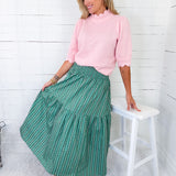 Woman wearing a pink top and green skirt sitting on a white stool against a white wooden wall.