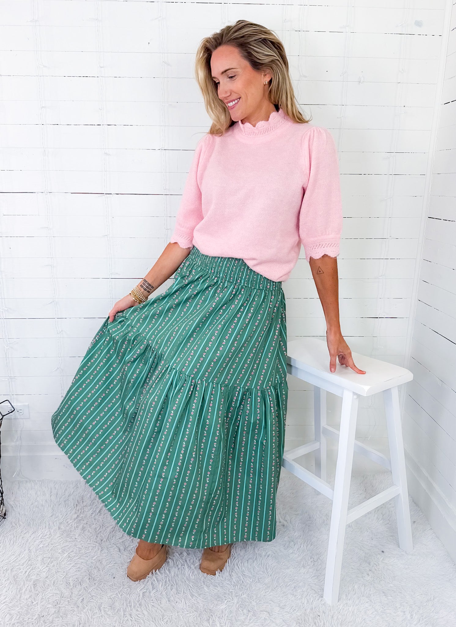 Woman wearing a pink top and green skirt sitting on a white stool against a white wooden wall.