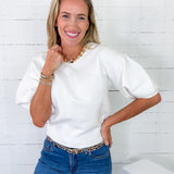 Woman wearing a white top and blue jeans with a gold belt, standing against a white wooden panel background.