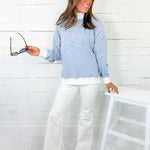 Woman wearing a light blue sweater and white pants in a room with a white chair and wooden floor.