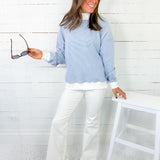 Woman wearing a light blue sweater and white pants in a room with a white chair and wooden floor.