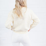 A woman with blonde hair is shown from behind, wearing a cream and white striped long-sleeve sweatshirt and white pants, standing against a white wood-paneled wall.