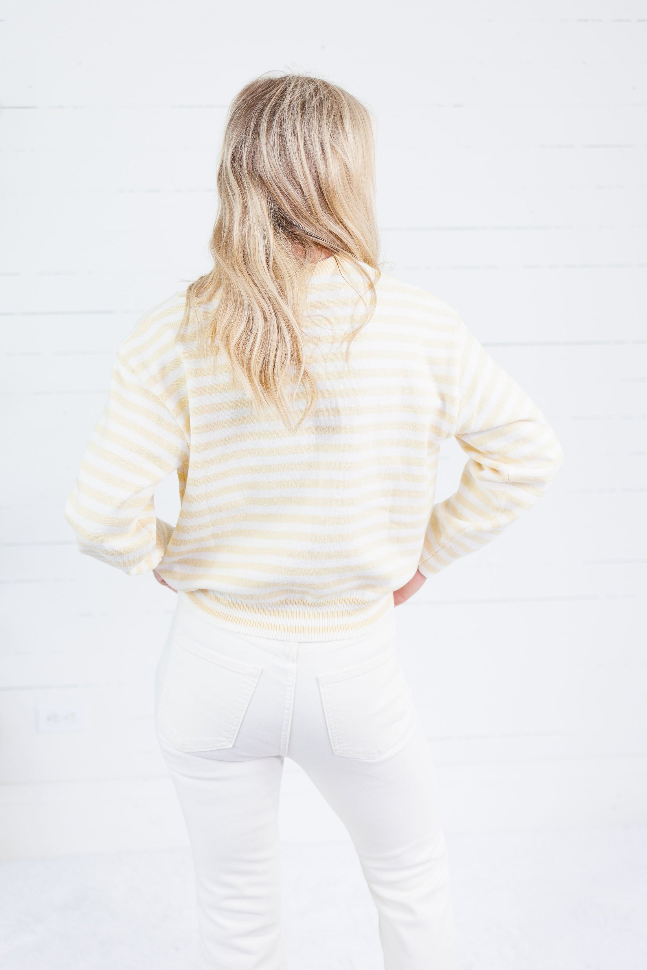 A woman with blonde hair is shown from behind, wearing a cream and white striped long-sleeve sweatshirt and white pants, standing against a white wood-paneled wall.