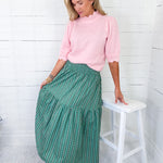 Woman wearing a pink sweater and green patterned skirt standing next to a white stool against a white wooden wall.