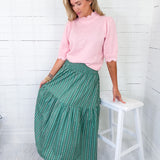 Woman wearing a pink sweater and green patterned skirt standing next to a white stool against a white wooden wall.