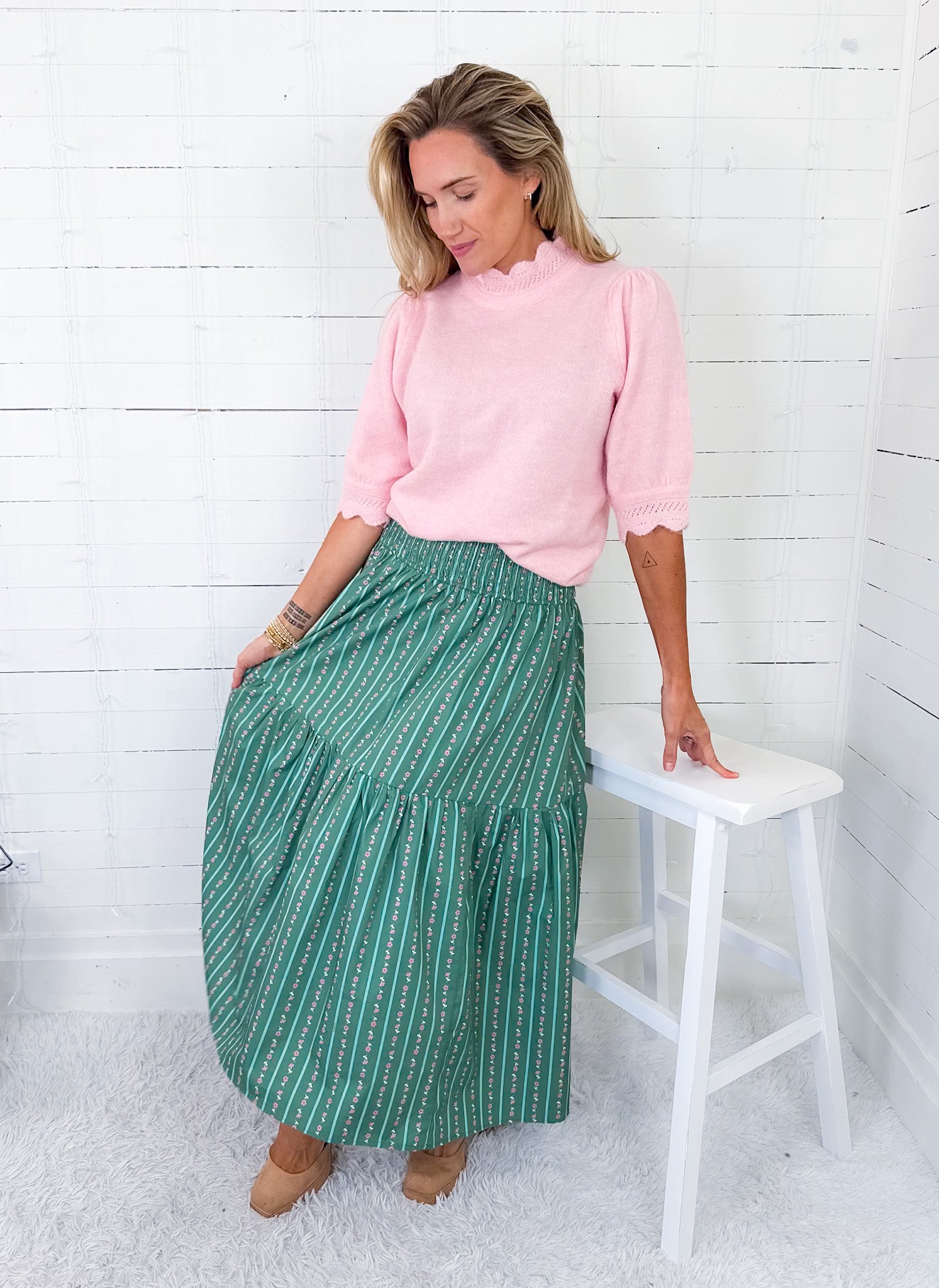 Woman wearing a pink sweater and green patterned skirt standing next to a white stool against a white wooden wall.
