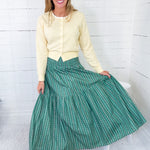 Woman wearing a yellow sweater and green skirt sitting on a white stool against a white wooden panel background.