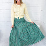 Woman wearing a yellow sweater and green skirt sitting on a white stool against a white wooden panel background.