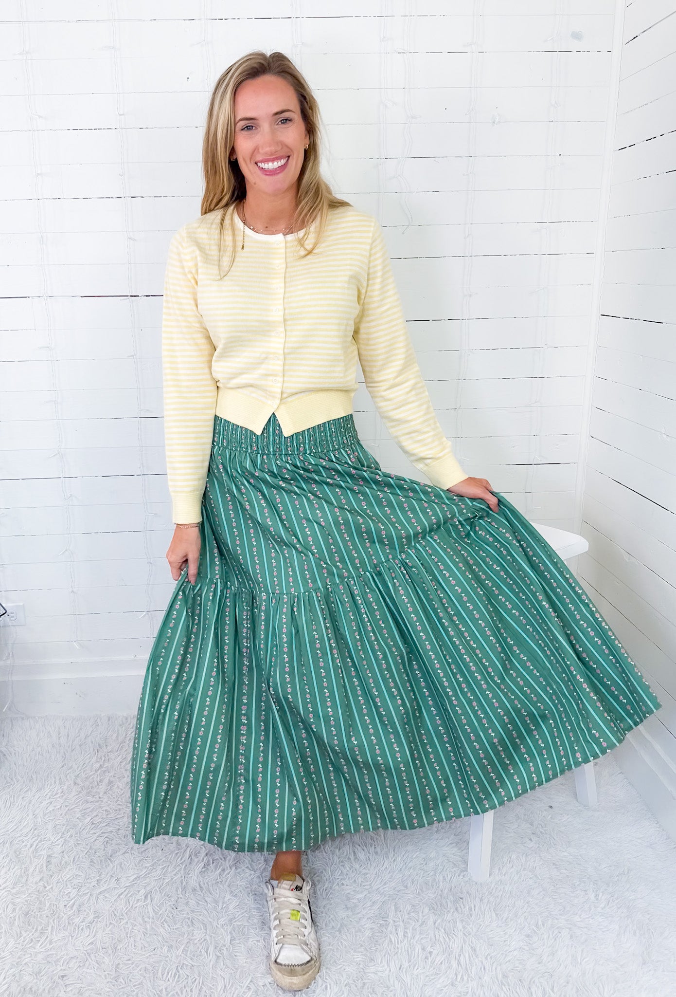 Woman wearing a yellow sweater and green skirt sitting on a white stool against a white wooden panel background.