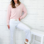 Woman wearing a pink sweater and white pants sitting on a white stool against a white wooden panel wall.