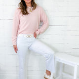Woman wearing a pink sweater and white pants sitting on a white stool against a white wooden panel wall.