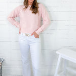 Woman wearing a pink sweater and white jeans standing against a white wooden panel wall.