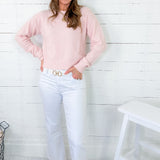 Woman wearing a pink sweater and white jeans standing against a white wooden panel wall.